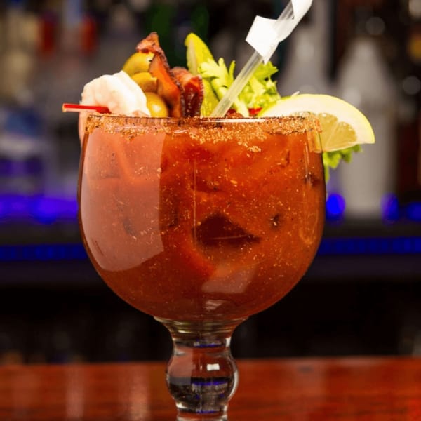 A Bloody Mary cocktail in a goblet, garnished with bacon, shrimp, olives, celery, and a lemon slice.