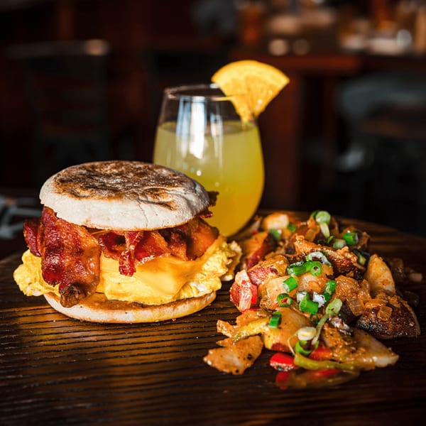 A breakfast sandwich with bacon, egg, and cheese, served with home fries and a mimosa.