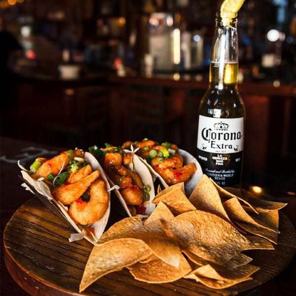 Three shrimp tacos with chips and a Corona beer bottle on a wooden board at Village Idiot Pub.