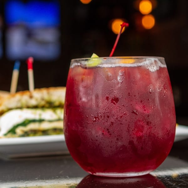 A vibrant red cocktail with a lime wedge and cherry, next to a sandwich on a plate.