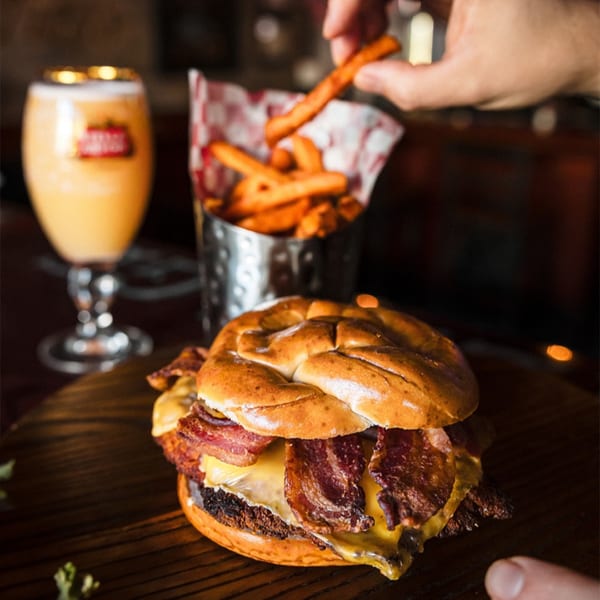 A Village Idiot Pub bacon cheeseburger with sweet potato fries and a Stella Artois beer.